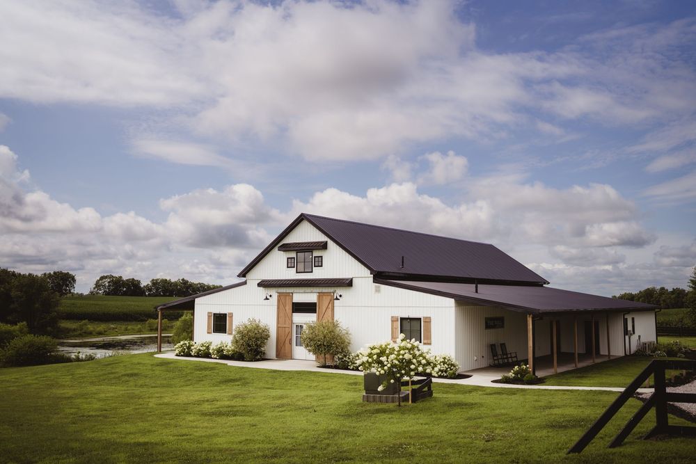 Modern white farmhouse with multiple buildings and structures sprawled across green rolling hills under a cloudy blue sky.