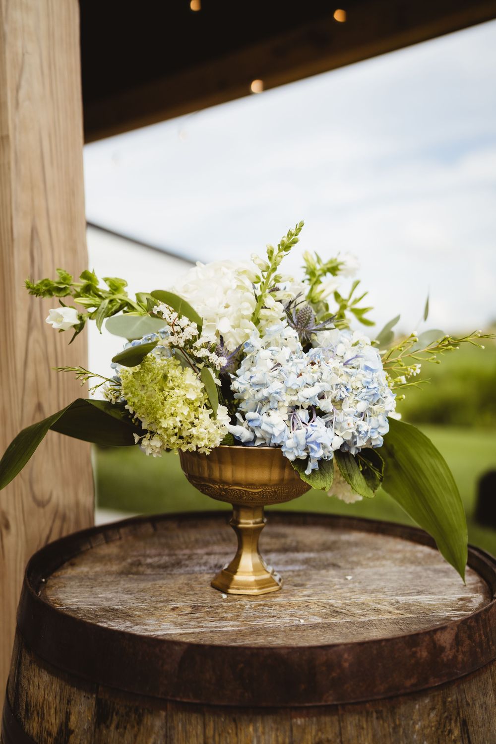 White and blue hydrangeas arranged in a gold pedestal vase sit atop a rustic wooden barrel outdoors.