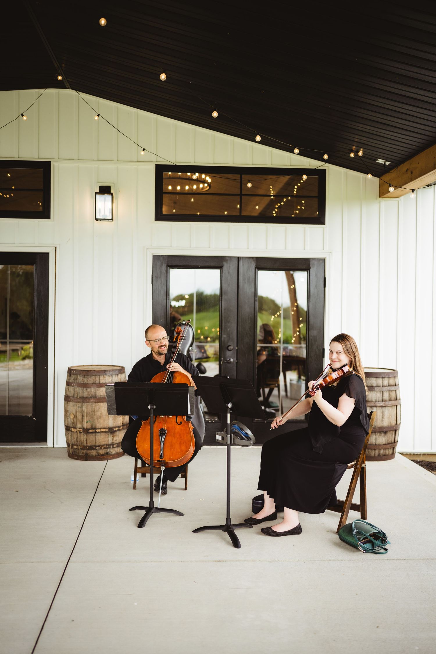 Musicians perform on cello and violin at a modern barn venue with rustic barrels and string lights overhead.
