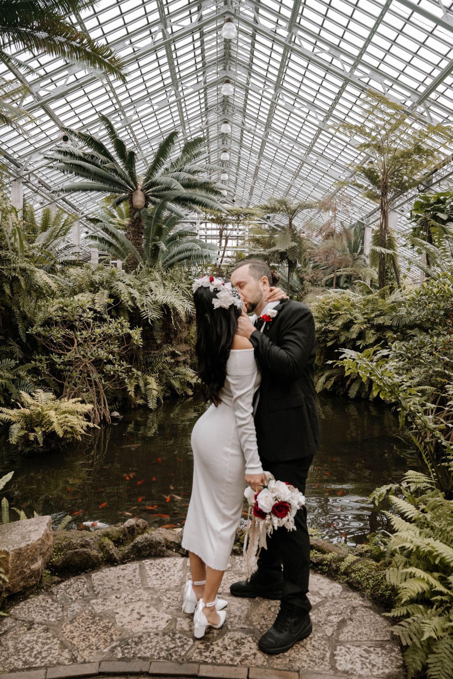 A romantic greenhouse wedding ceremony takes place among lush palm trees and tropical plants under a glass ceiling.