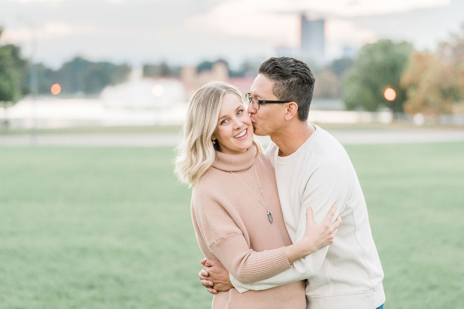 A romantic couple shares tender moments in a soft-lit meadow wearing blush and white outfits during an engagement photoshoot.