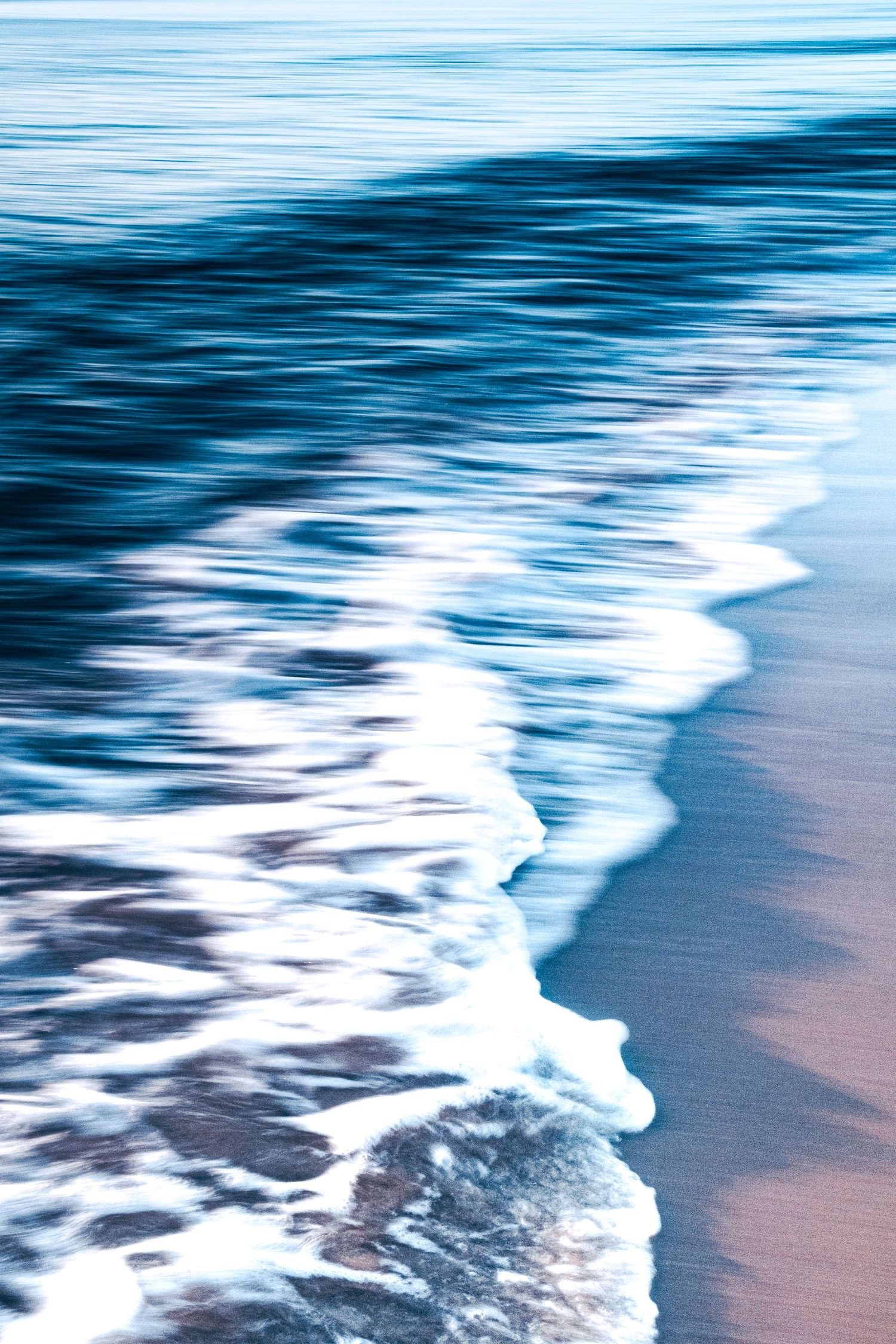 Long exposure blue-toned photo of a soft ocean wave gently washing onto a sandy beach shoreline.