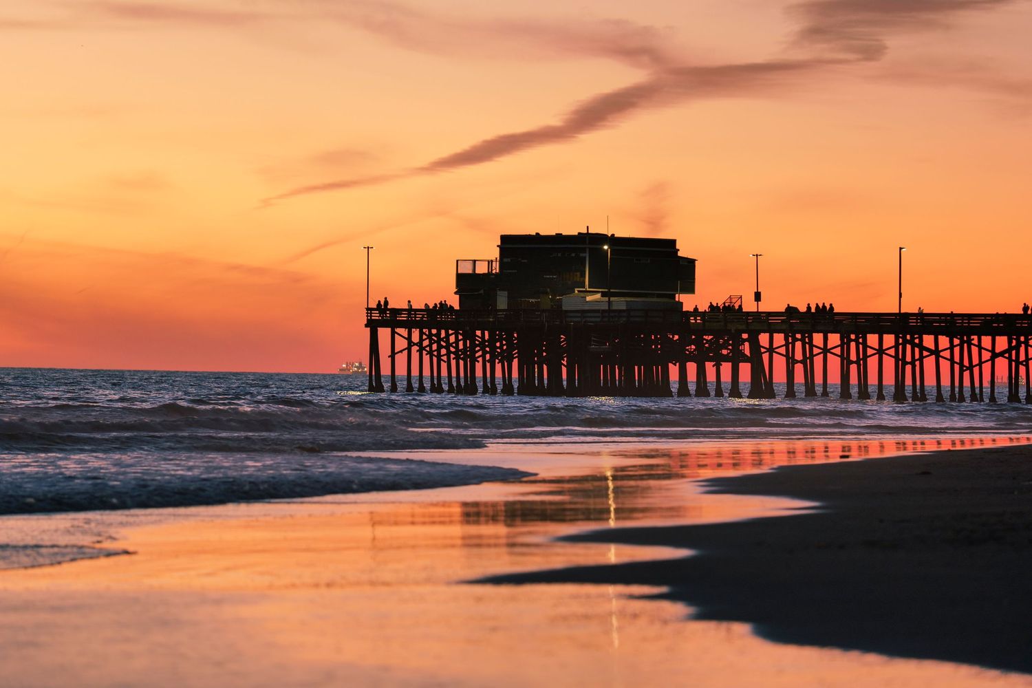 Silhouette of a beach pier reflected in wet sand during a vibrant orange and red coastal sunset.