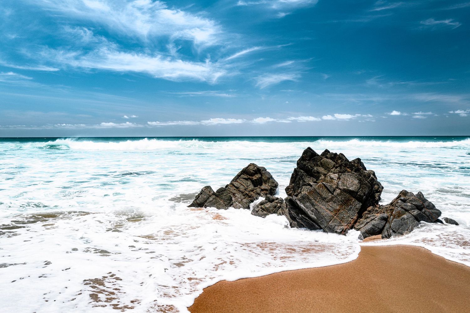 Turquoise ocean waves crashing around dark coastal rocks on a sandy beach under blue sky.