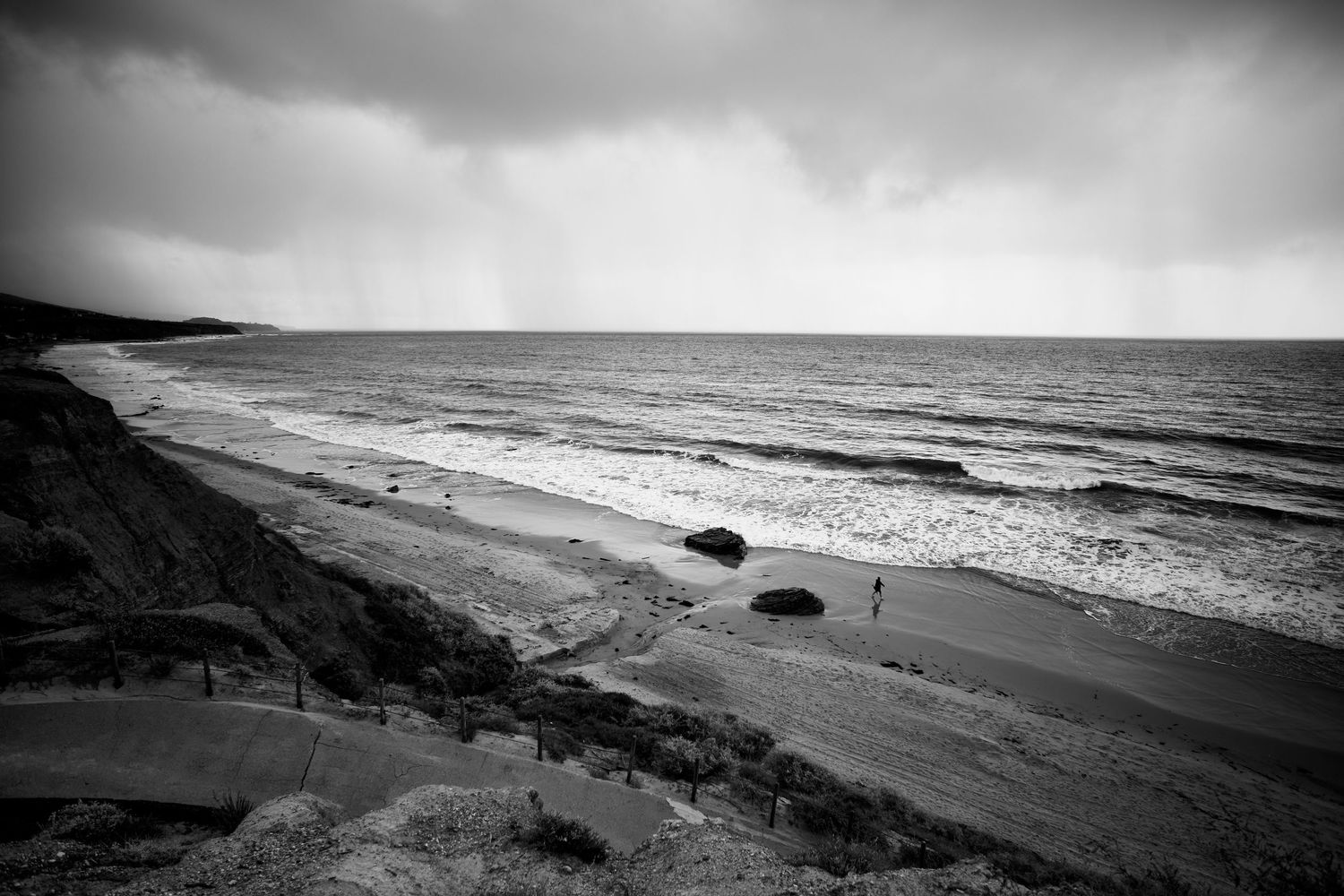 Black and white stormy beach scene with rough waves, sandy cliffs, and distant rain.