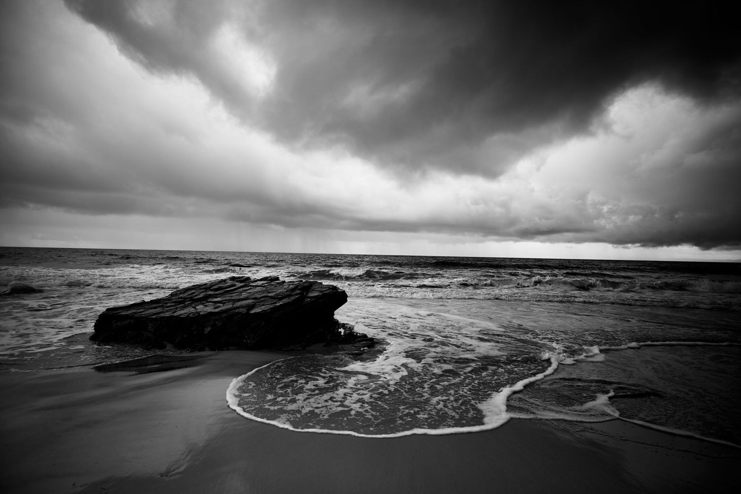 Dramatic black and white seascape with a lone rock surrounded by ocean foam under stormy clouds.