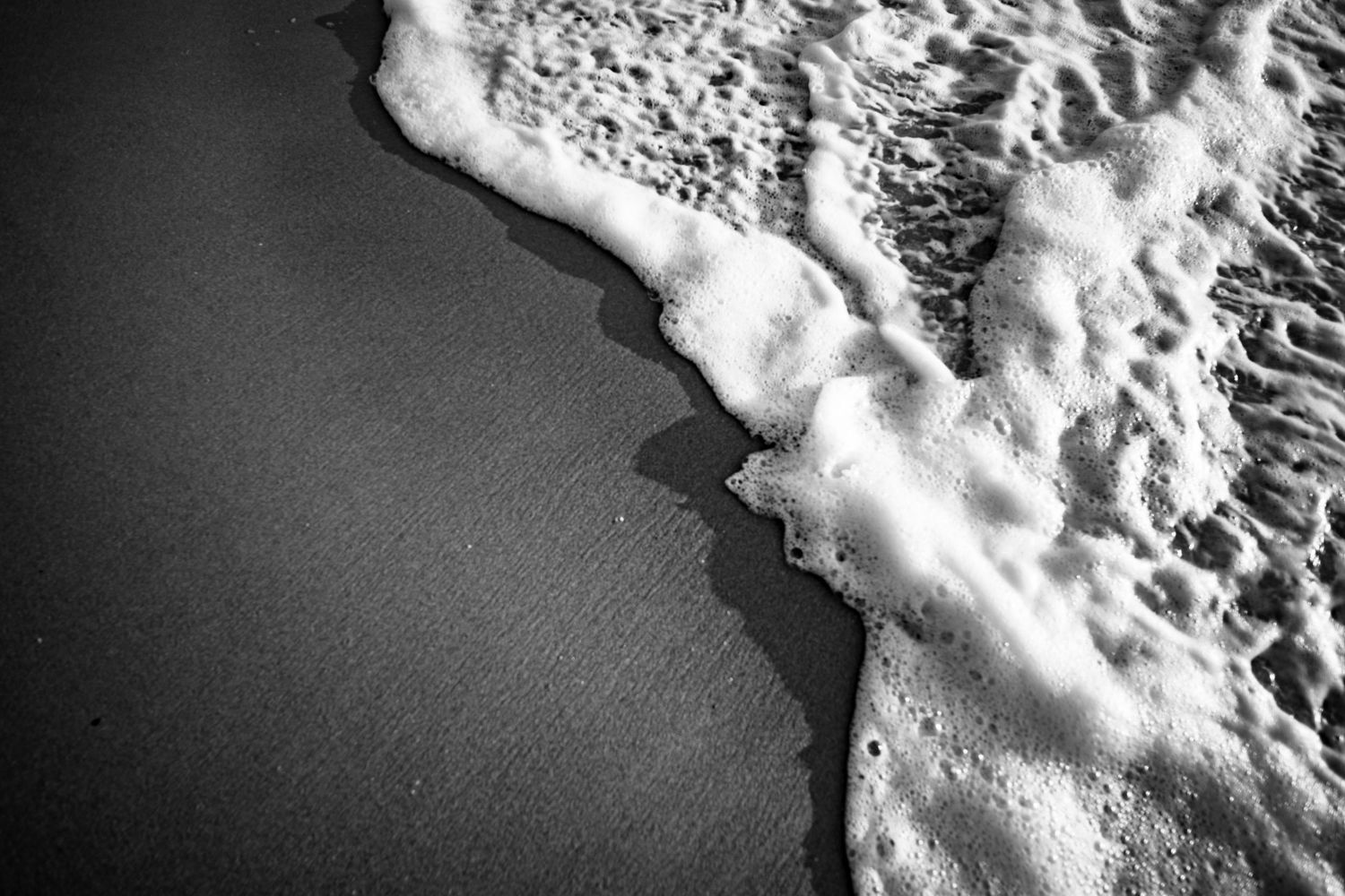 Black and white aerial view of a foamy ocean wave washing onto a smooth dark sandy beach shore.