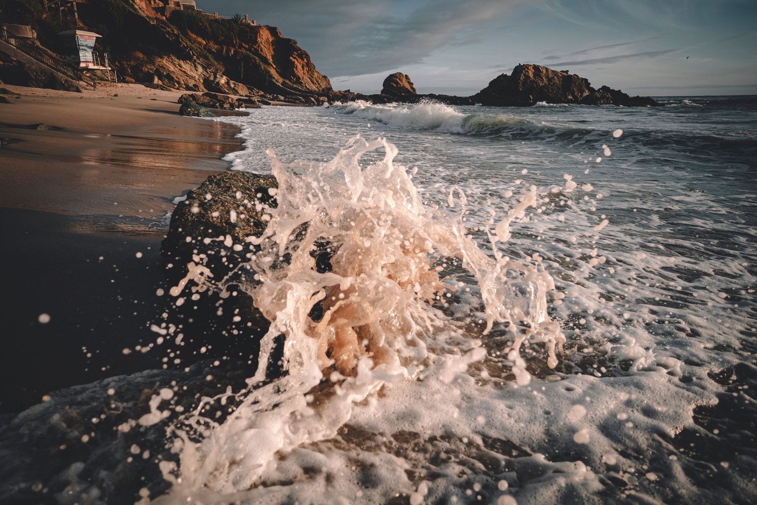 Dynamic ocean wave splashing against rocks on a scenic California beach with dramatic cliffs in golden light.