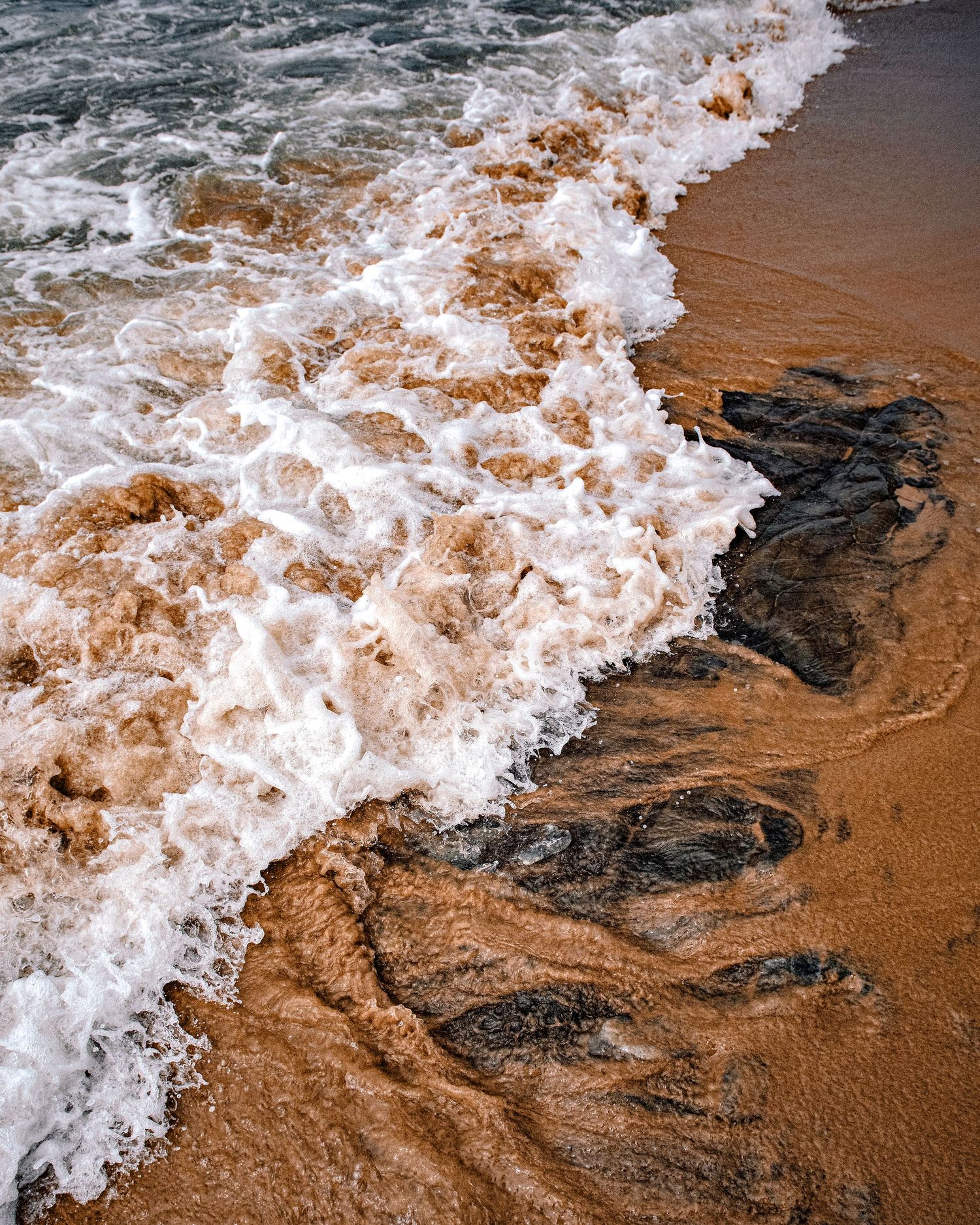 Aerial view of turbulent brown foamy ocean waves washing over dark wet sand on a beach shoreline.