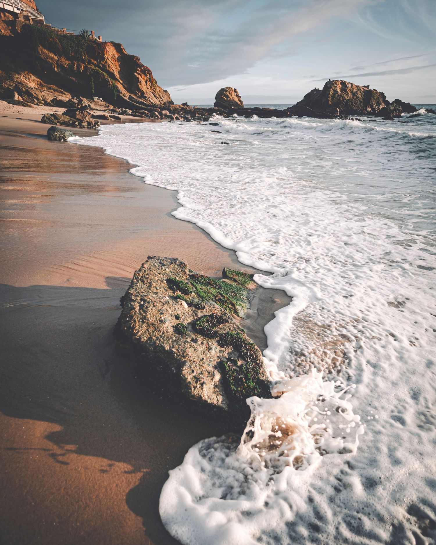 Foamy ocean waves washing over a mossy rock on a sandy beach with rocky cliffs in warm sunlight.