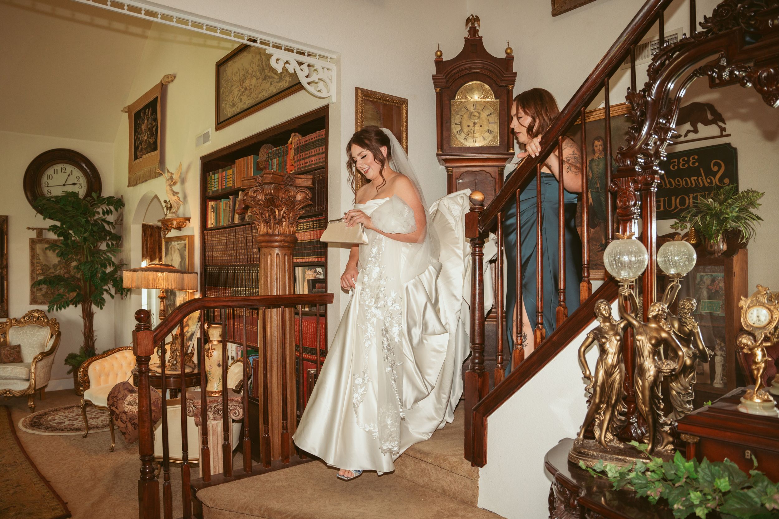 An elegant bride descends a vintage wooden staircase surrounded by antique decor and bookshelves.