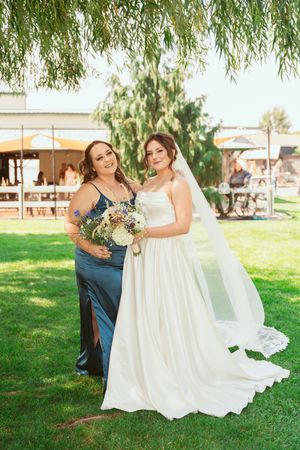 A bride in a flowing white gown poses with a bridesmaid in a navy blue dress under a willow tree at an outdoor venue.