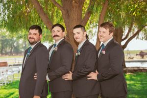 Groomsmen in black suits pose together under trees at an outdoor wedding venue.
