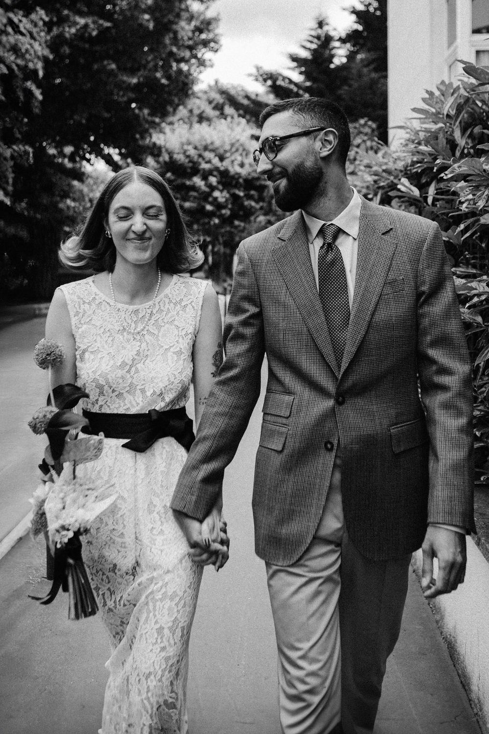 Black and white photo of smiling couple walking hand in hand, bride in lace dress, groom in checked suit.