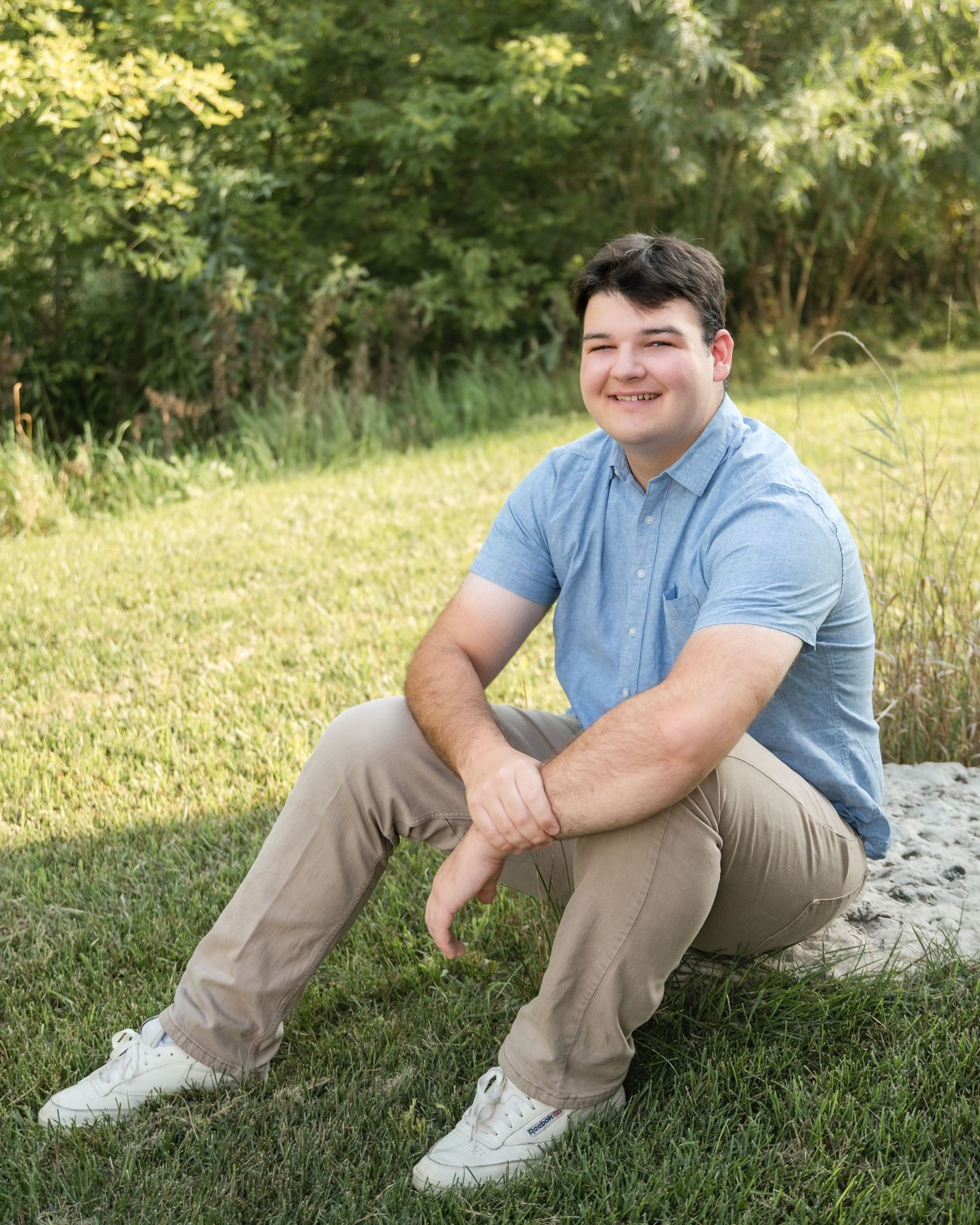 A person in a light blue shirt and khaki pants sits casually on grassy ground during a sunny outdoor portrait session.