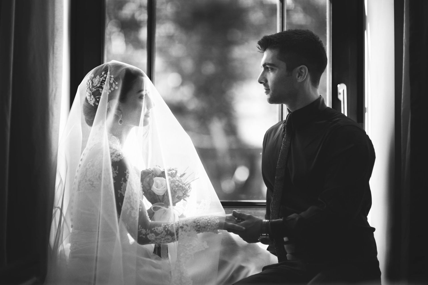 Black and white silhouette of bride and groom facing each other near window in romantic moment during wedding.