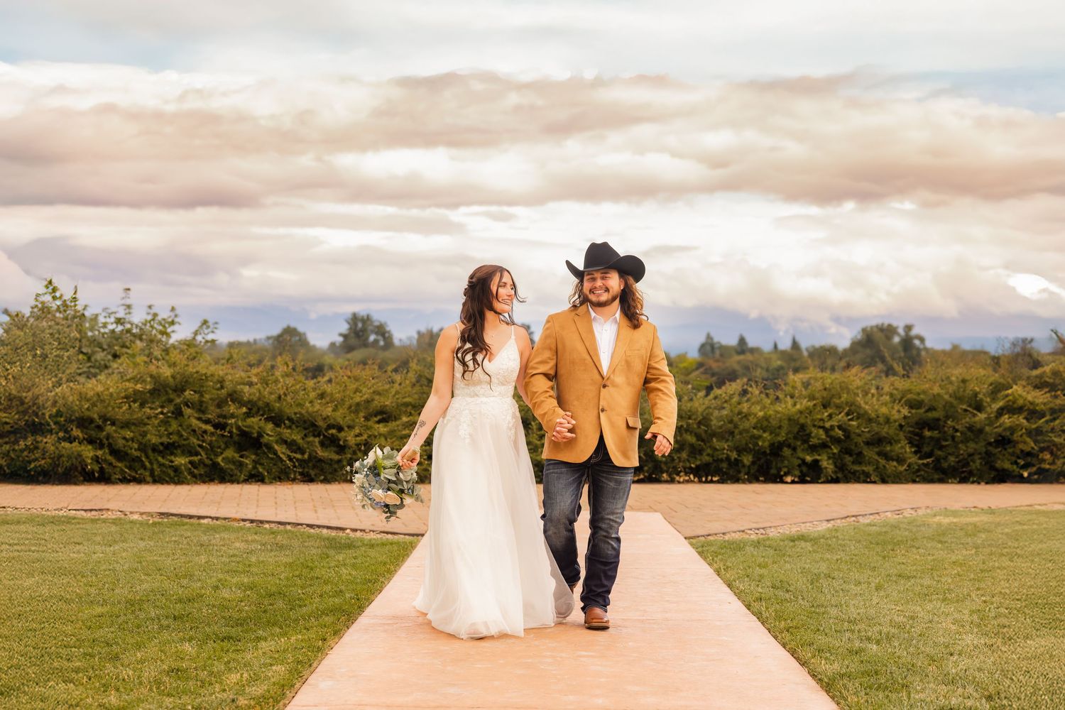 A couple walks hand in hand on a path surrounded by rolling hills and dramatic clouds at sunset.