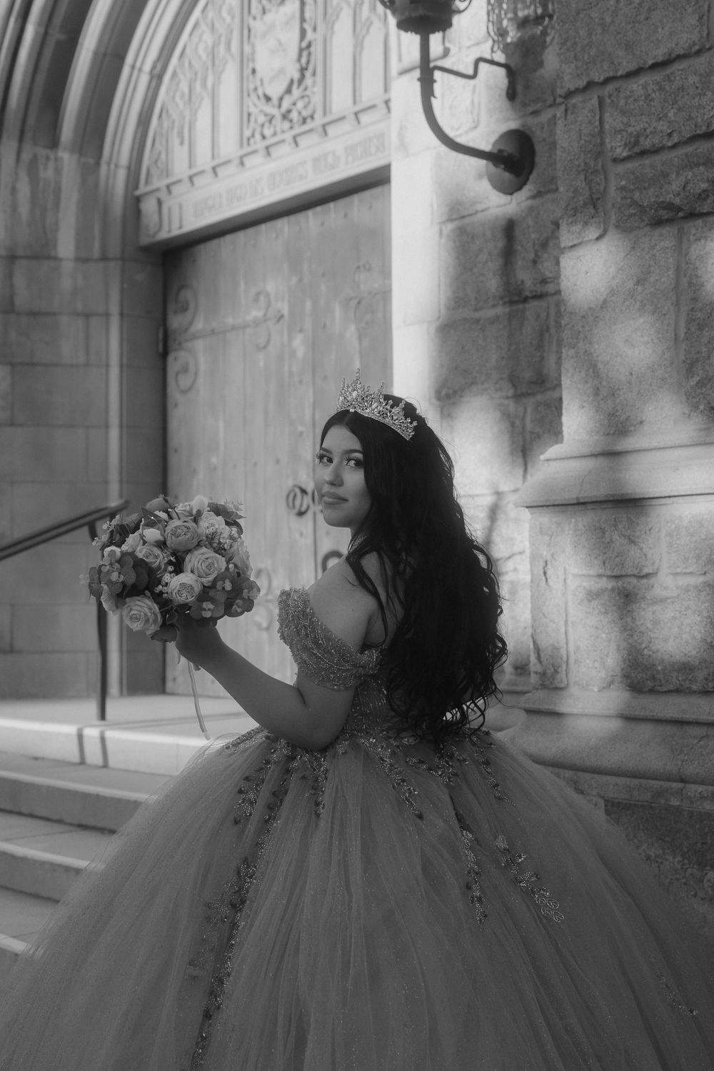 A figure in an elegant ballgown and tiara poses with a bouquet against ornate stone architecture in black and white.