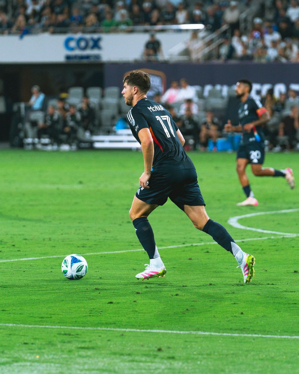 A man dribbles a soccer ball on the field with a full crowd behind him