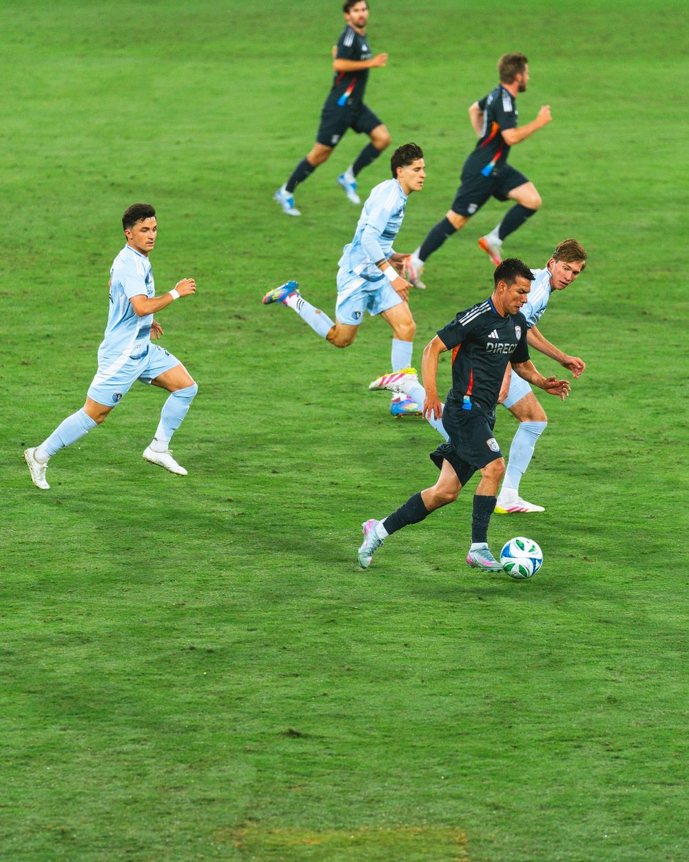 Several men play soccer on a green field, one person dribbles the ball