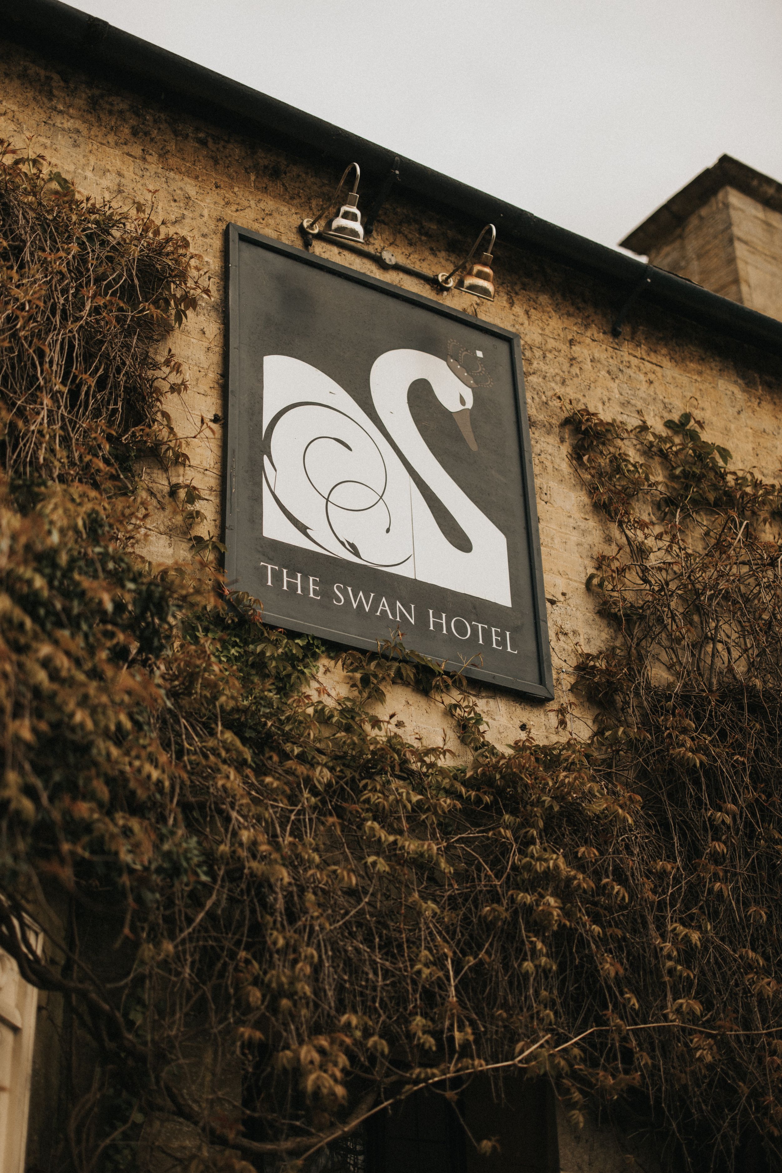 A black and white sign for The Swan Hotel features an elegant swan logo on a weathered stone building covered in vines.