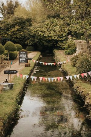 Colorful bunting flags hang over a tranquil garden stream with decorative signage and manicured hedges along the banks.