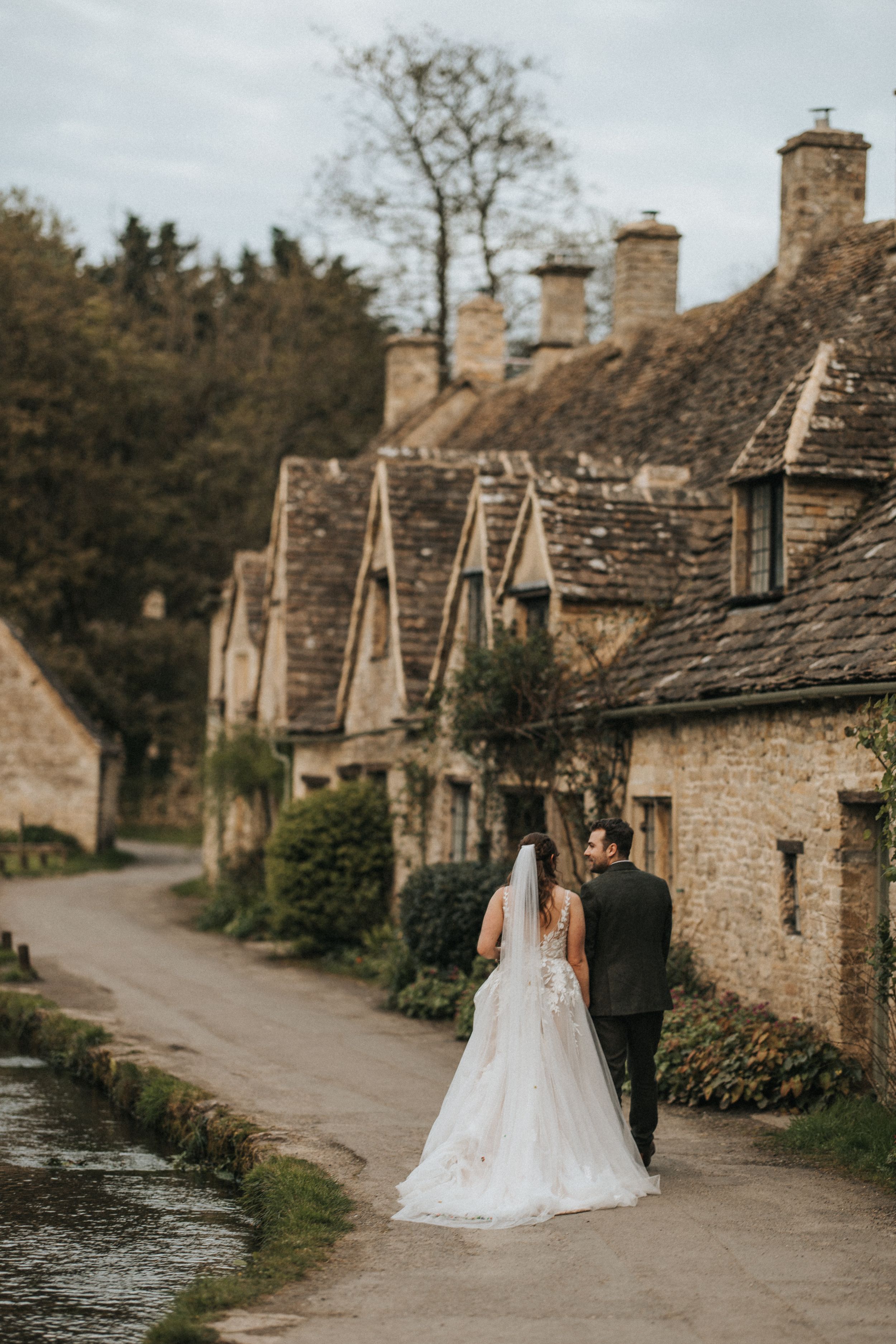 A couple in wedding attire walking along a scenic path beside stone cottages with mossy roofs in the Cotswolds.