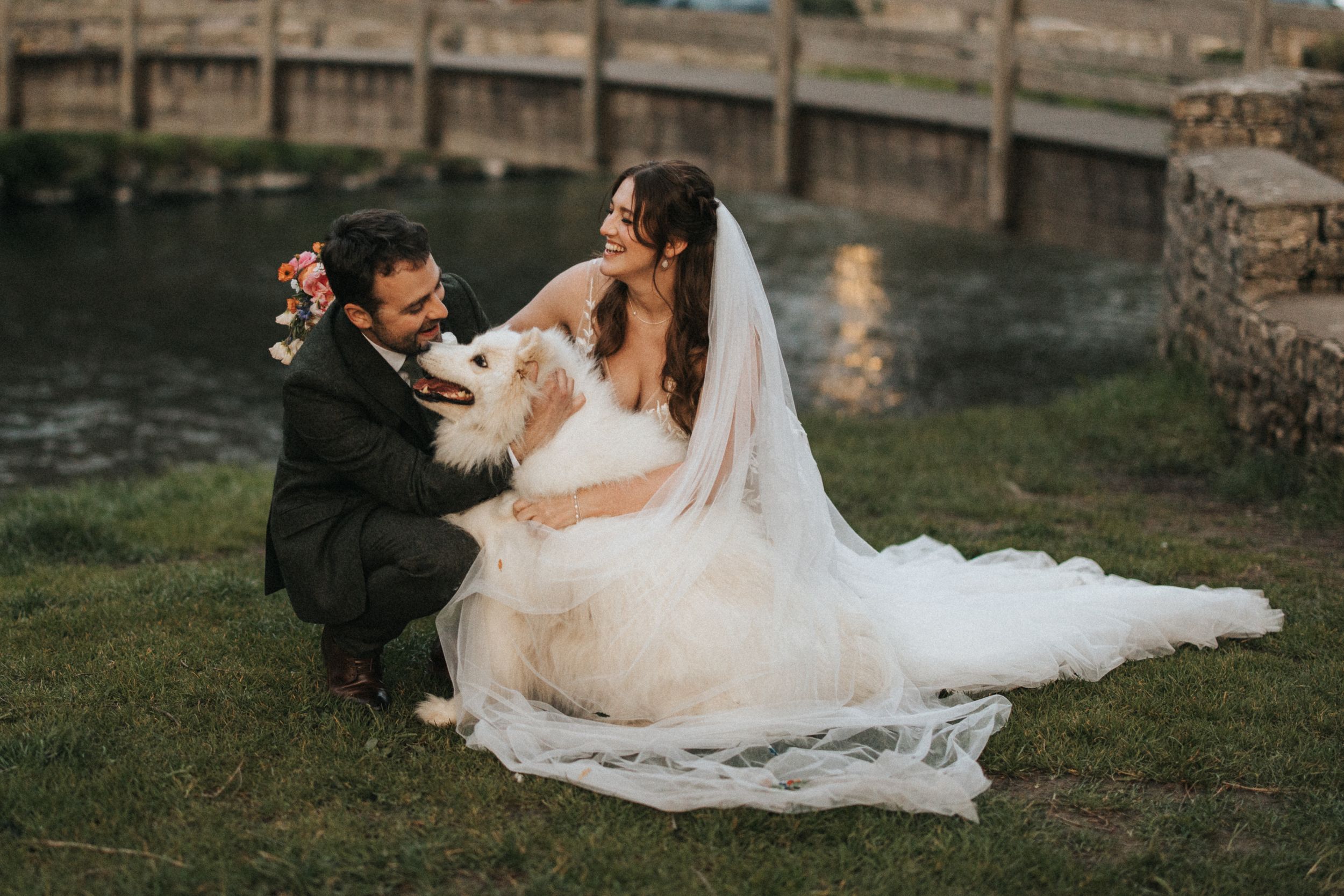 Two people share an intimate moment on grassy waterfront during sunset wedding photoshoot.