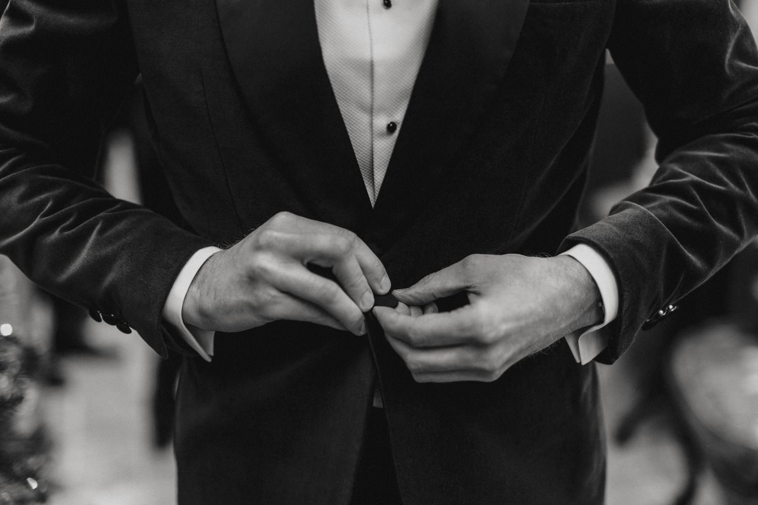 Black and white close up of hands adjusting buttons on a formal tuxedo jacket.