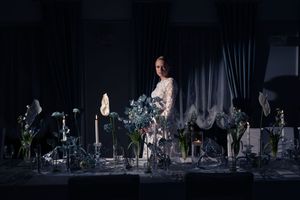 A person stands on stage amid dramatic floral arrangements and candlelight in an elegant theatrical setting.