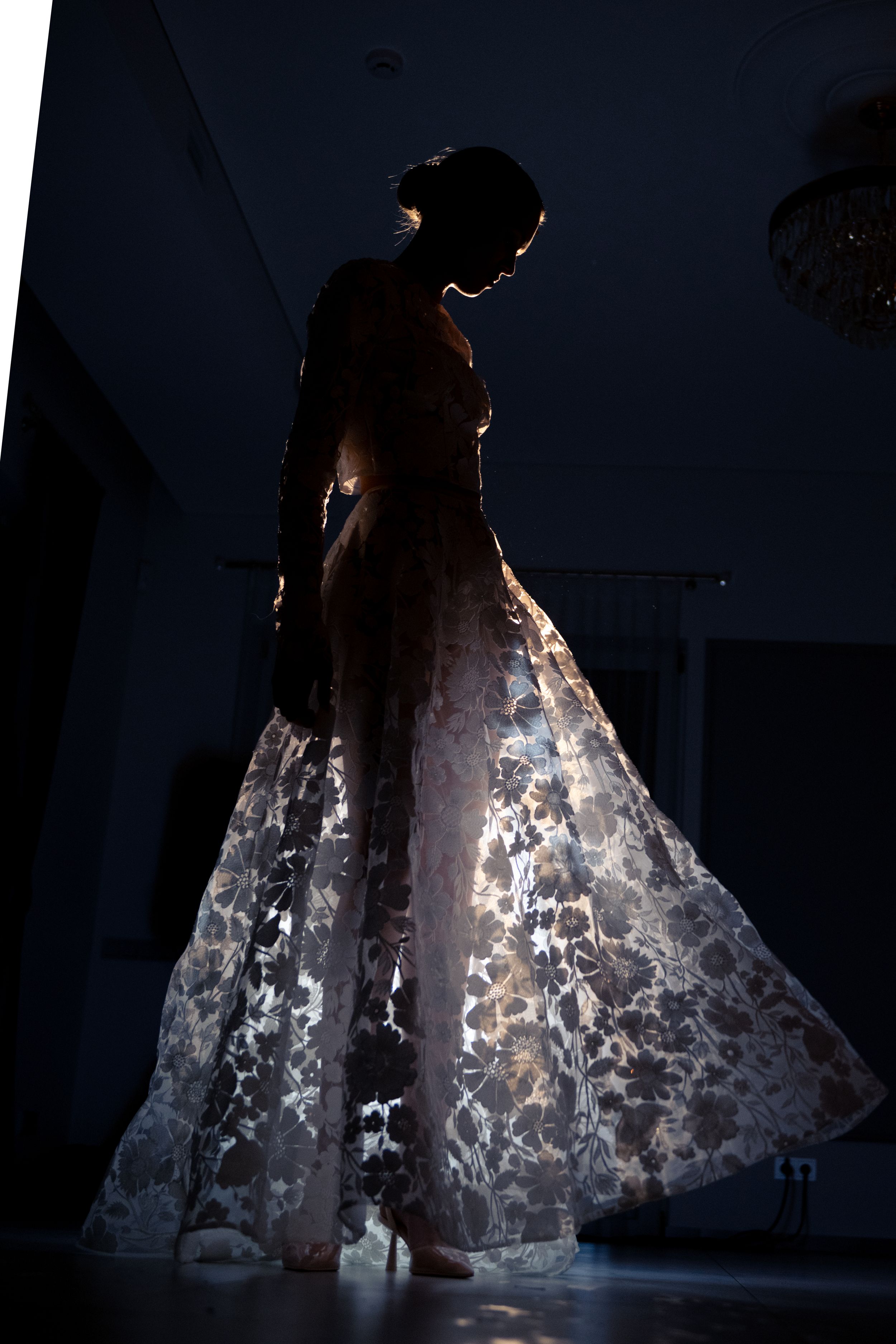 A silhouette in a glowing white lace wedding gown stands elegantly backlit in a dark room.