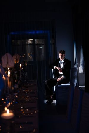 A dark atmospheric scene with candles illuminating a dimly lit room while someone sits at a table reading.