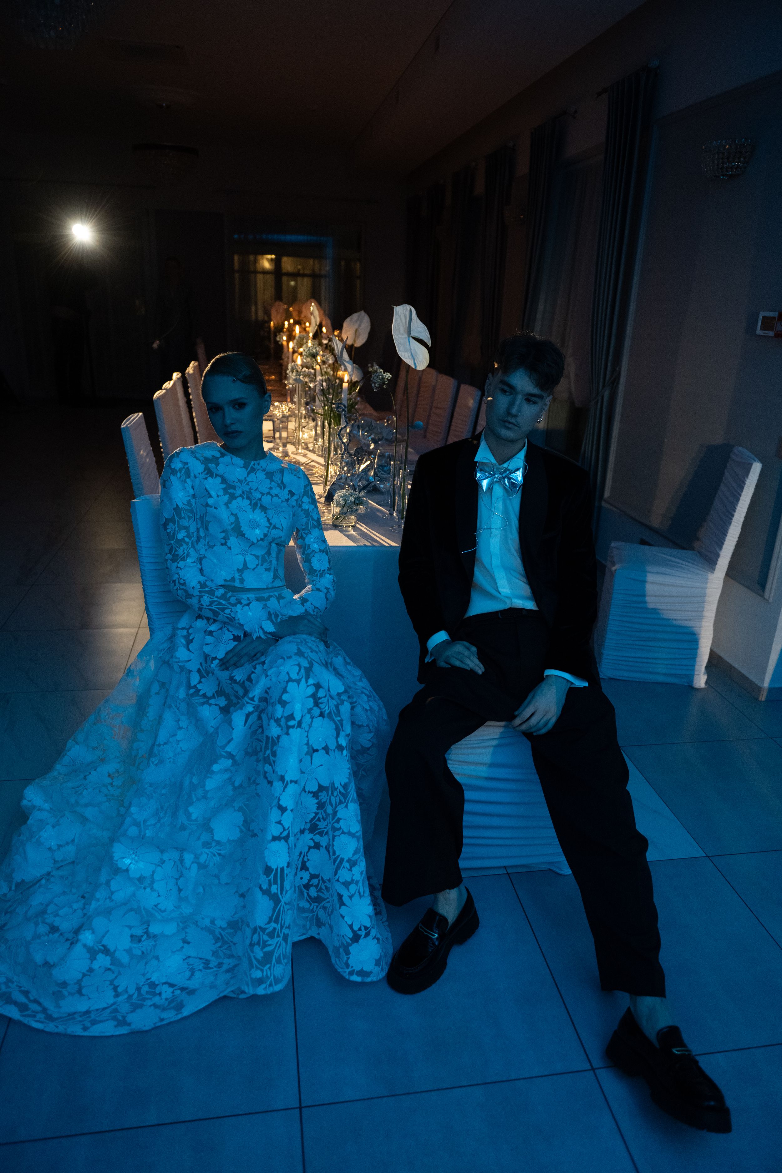 Couple in formal attire sits at an elegant dinner table illuminated by blue lighting and candlelight.