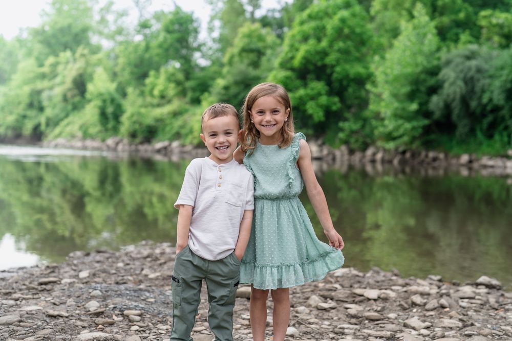 Two children in summer outfits hold hands while standing next to a tranquil river on a sunny day at Brecksville Reserve Family Portrait Sesh