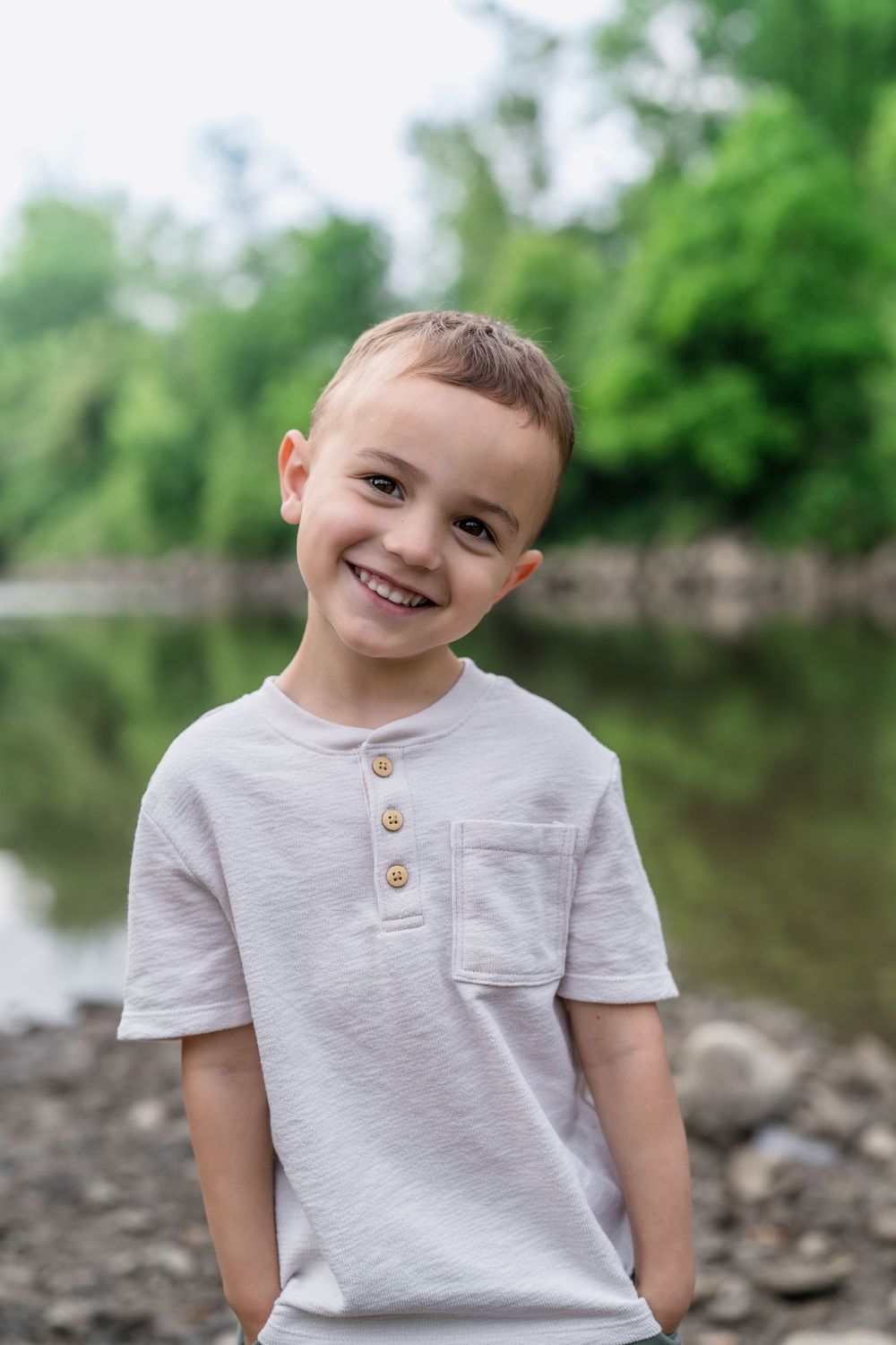 Young child wearing a gray t-shirt with button details poses outdoors on a rocky shoreline at Brecksville Reserve Family Lifestyle Session