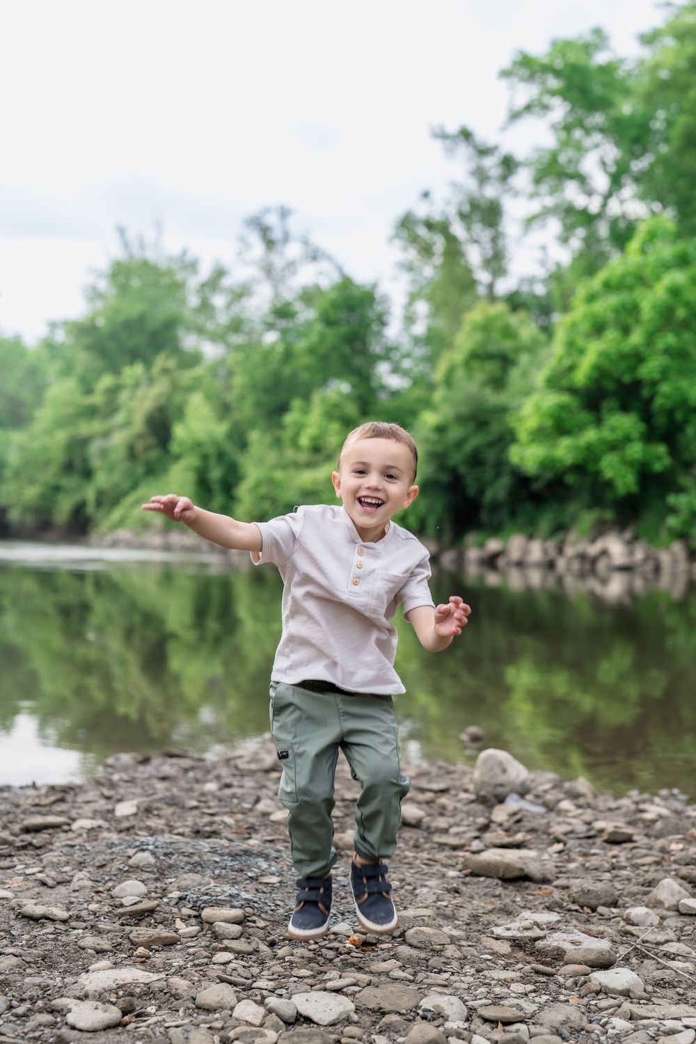 Child playfully leaping across rocky riverbank with trees and water from Brecksville Reserve in background of Family Session