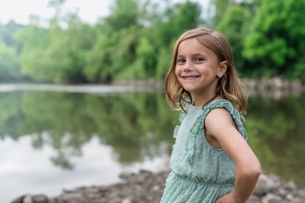 A young person in a mint green dress smiles by a serene lake with green trees in the background at Brecksville Reserve Family Photo Session