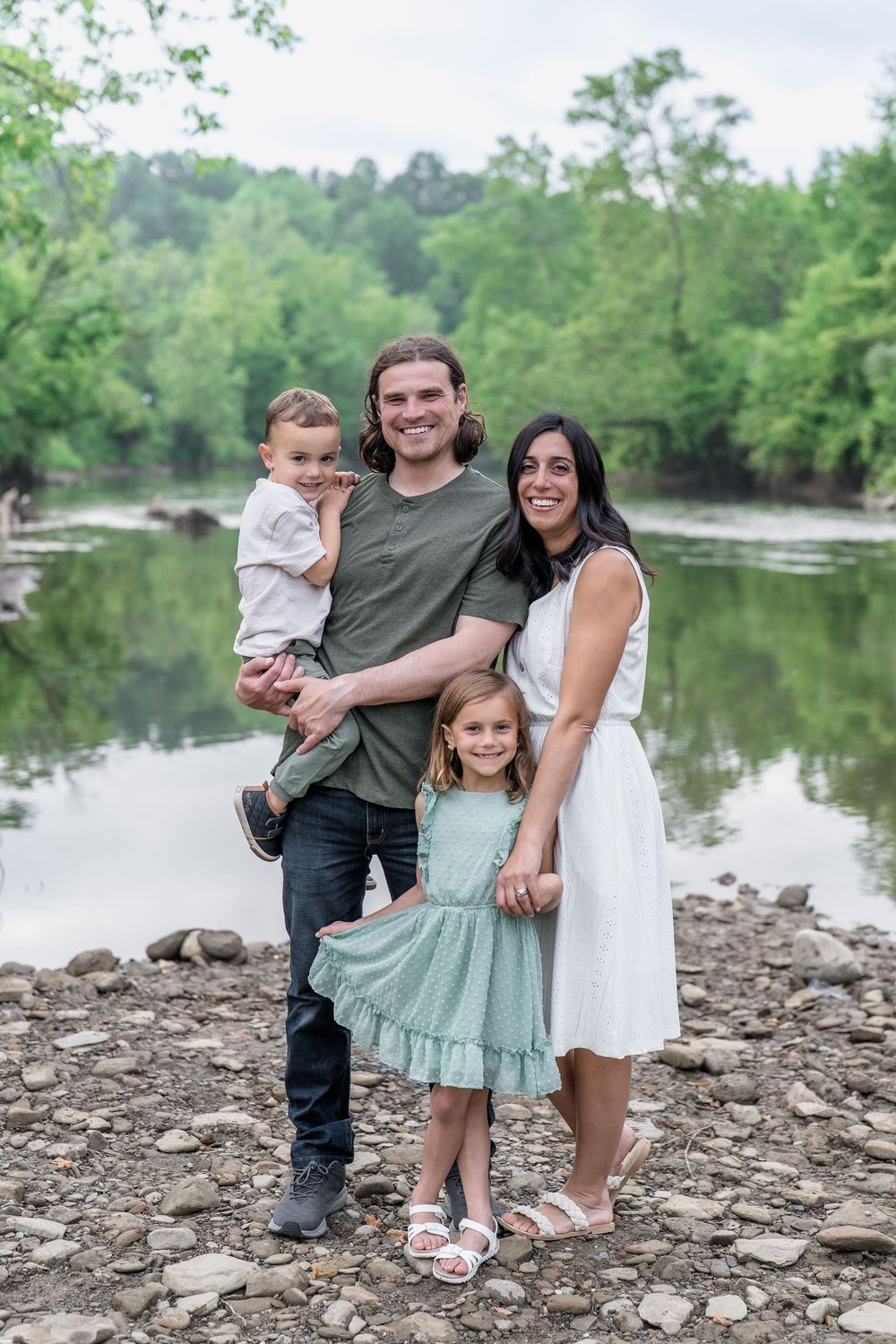 A family poses together on a rocky riverbank surrounded by green trees and calm water at Brecksville Reserve Family Photo Session.