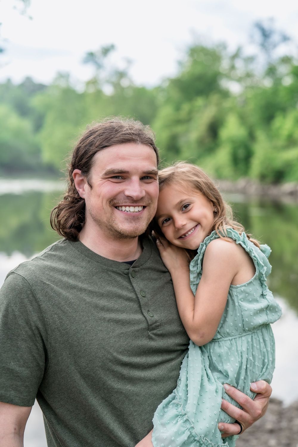 A parent and child share a tender moment outdoors by a lake, both smiling warmly in casual summer attire for Family Photo Session In Cle OH