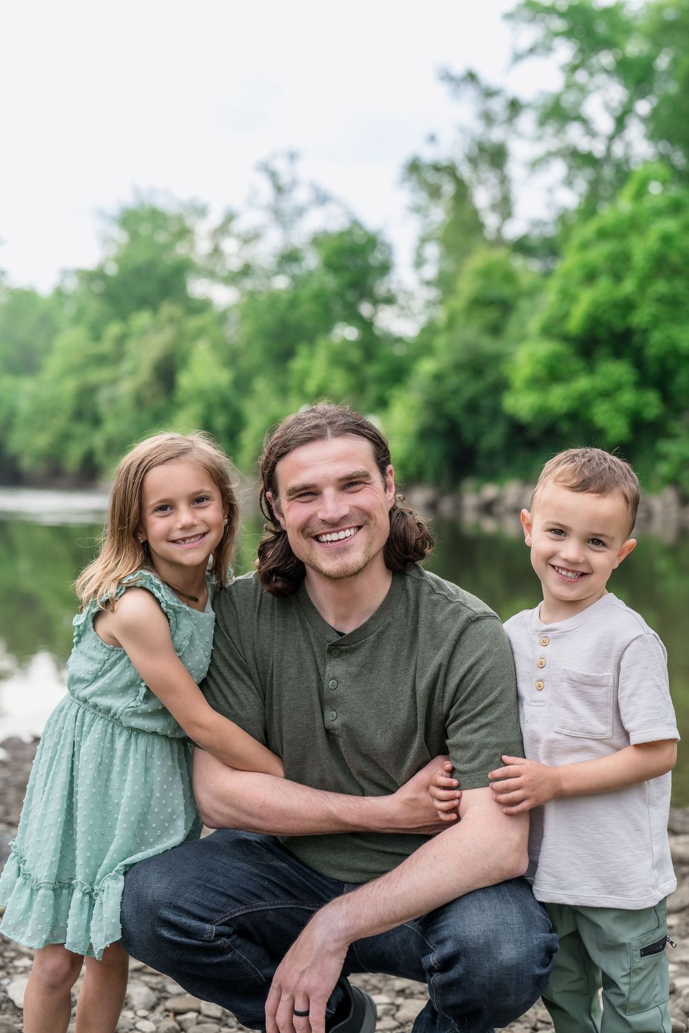 Family group sits together on rocky riverbank surrounded by lush green trees on a sunny day in Cleveland OH's family photo session
