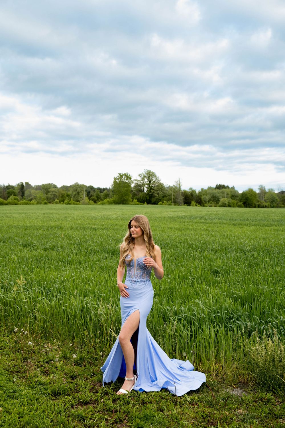 Elegant light blue evening gown with thigh slit photographed in lush green field under cloudy spring sky.