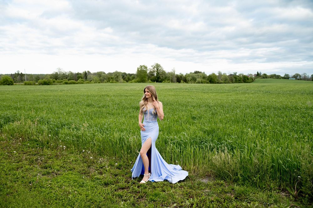 Long blue dress twirls against a lush green wheat field backdrop under cloudy skies.
