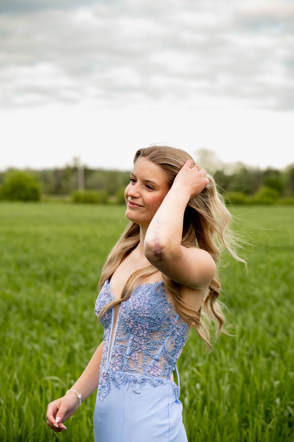 A person in a light blue embroidered top twirls joyfully in a lush green field under cloudy skies.