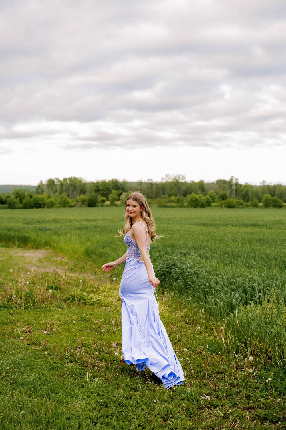 Woman in a flowing light blue gown twirls gracefully in a lush green meadow under cloudy skies.