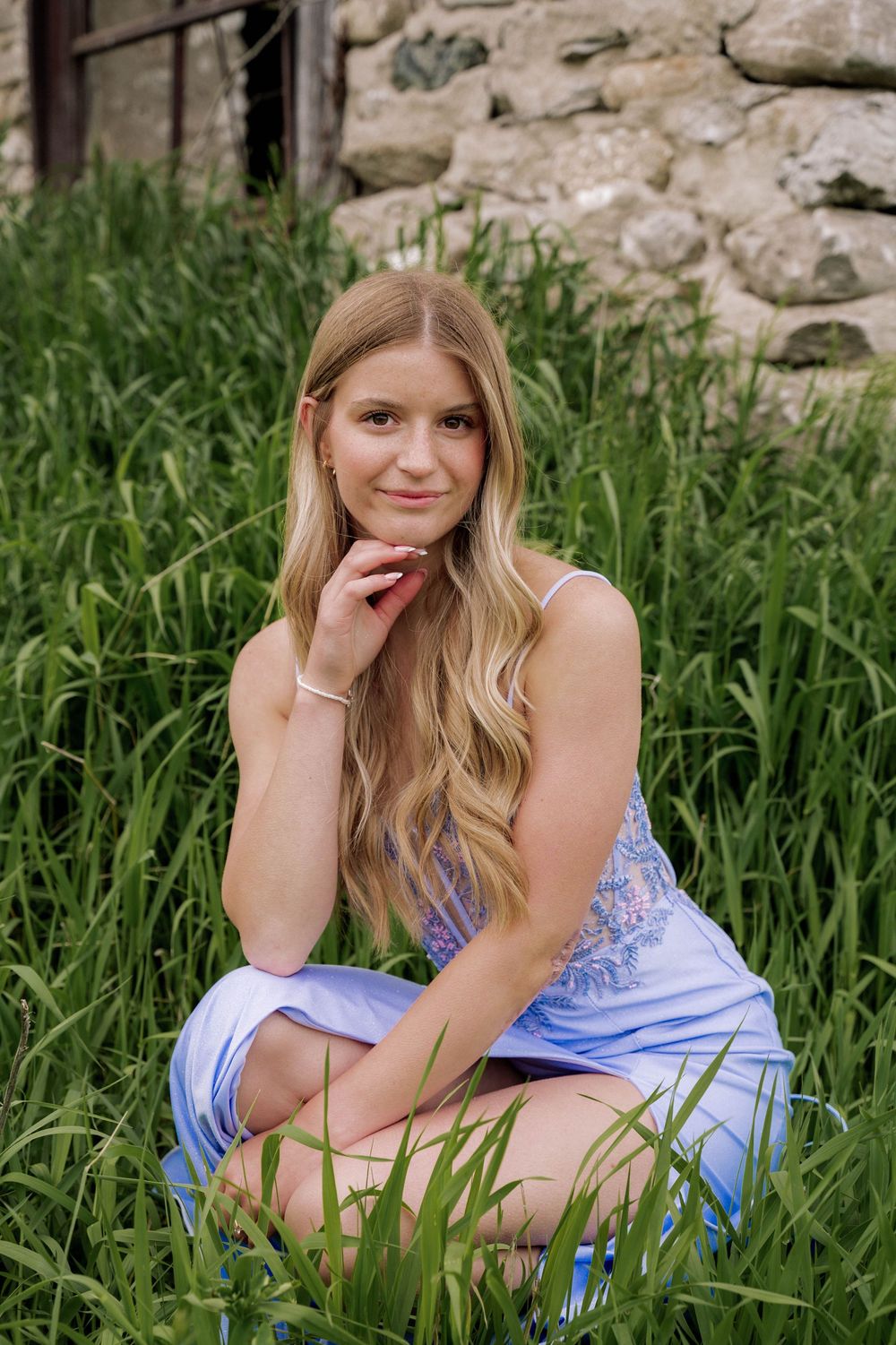 A portrait series of someone relaxing among tall grass near stone walls wearing a light blue off-shoulder top.