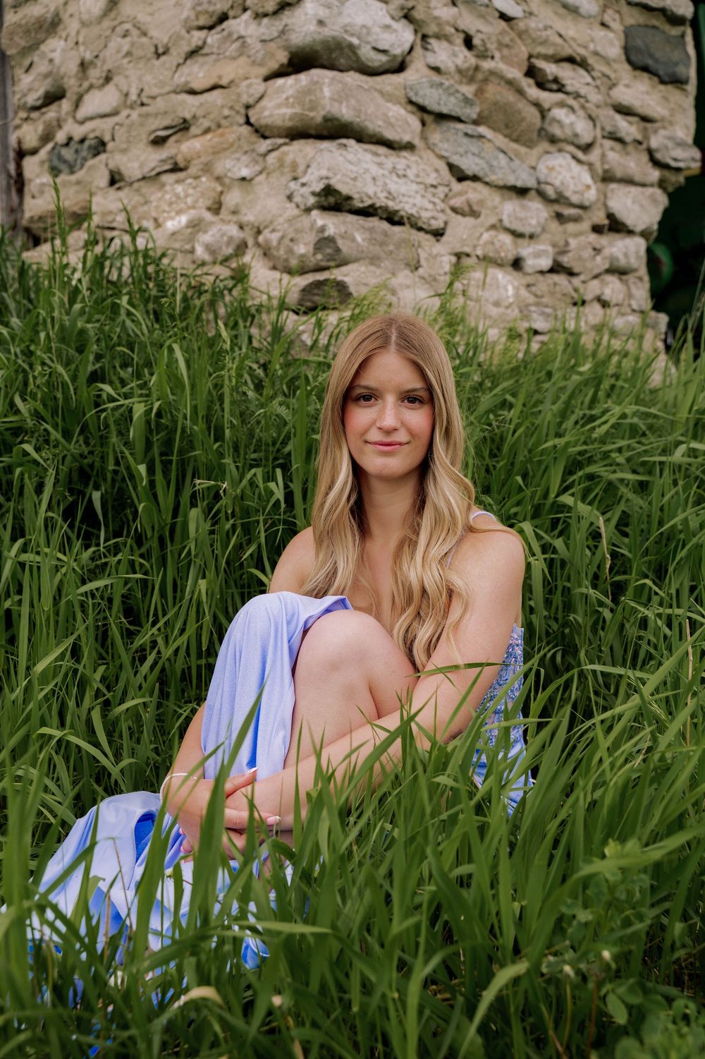 A figure sits in tall grass beside a rustic stone wall with purple irises blooming in a natural outdoor setting.