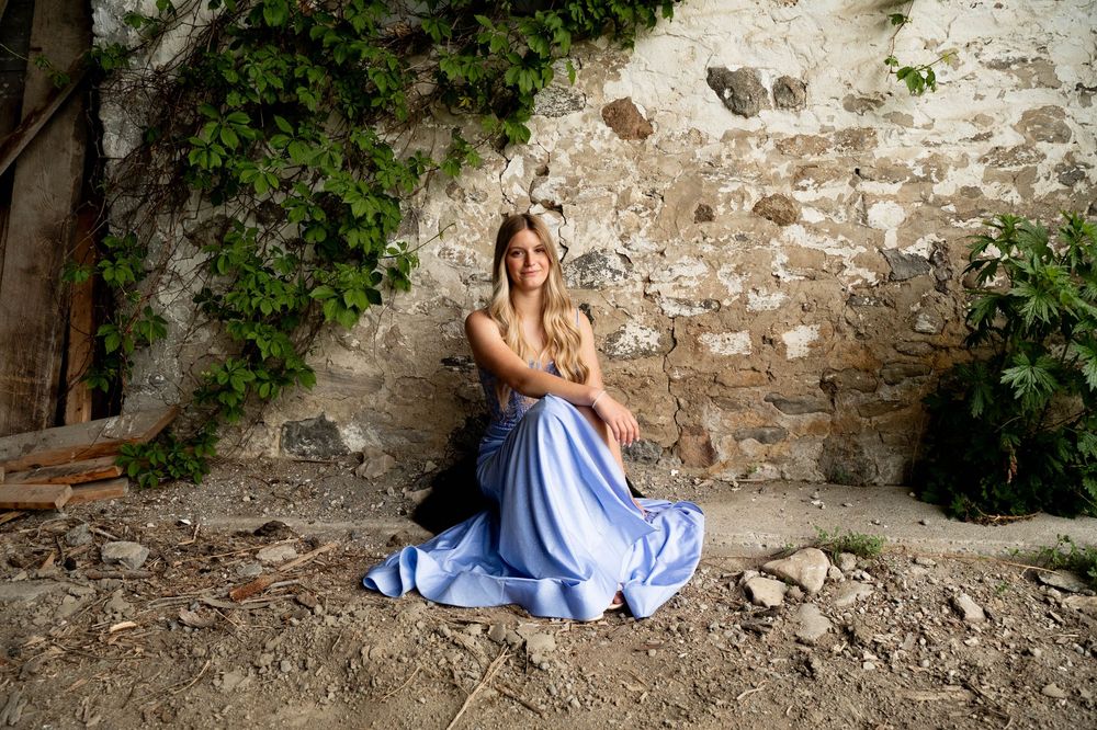 Person in a flowing blue dress sits against an old stone wall with climbing ivy in a rustic outdoor setting.