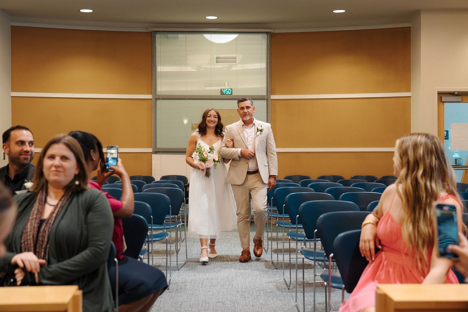 A couple proceeds down the aisle at an intimate indoor ceremony with blue chairs and guests looking on.
