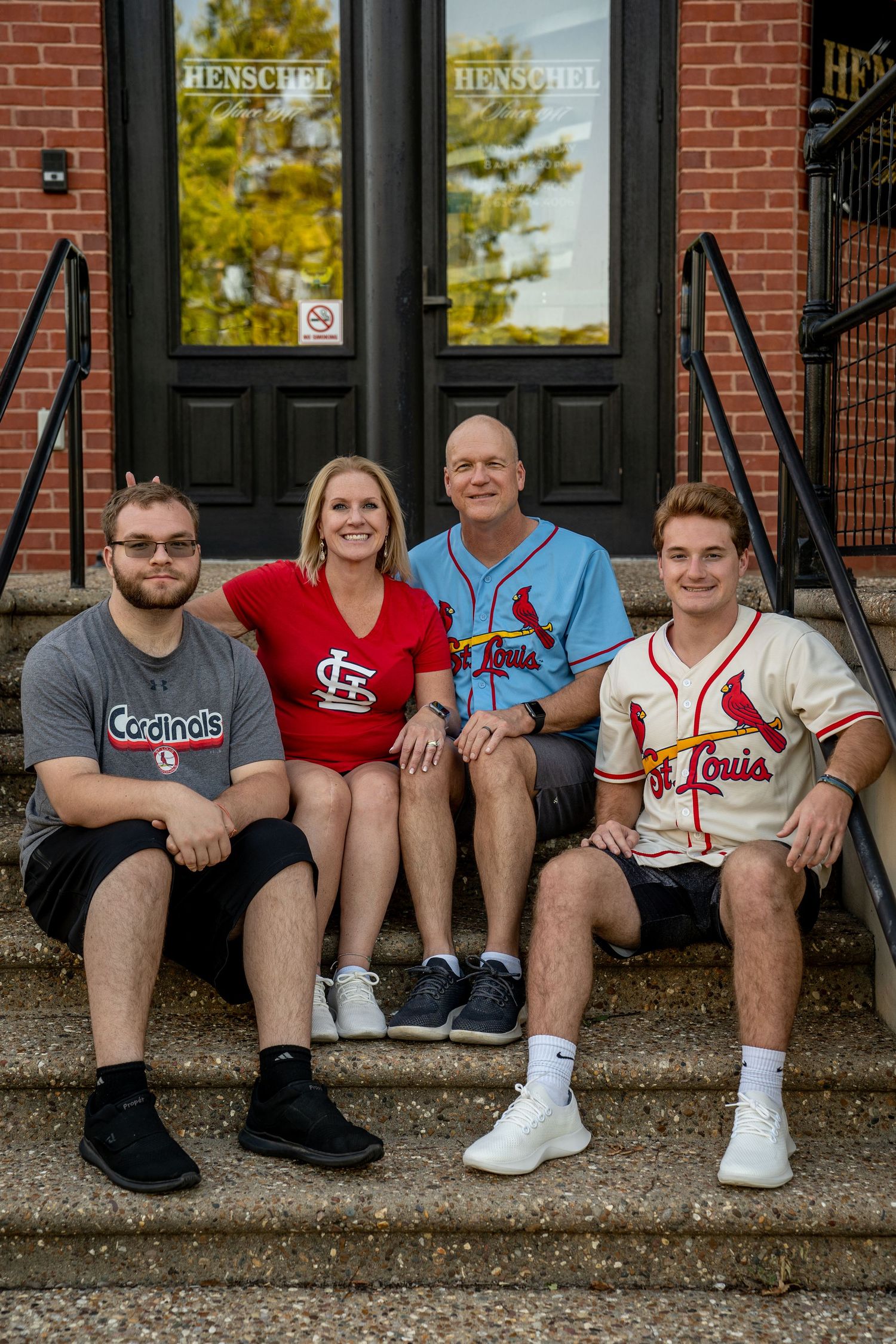 Family portrait of mom, dad and grown children wearing St. Louis jearseys sitting on steps leading up to a brick building with black doors.