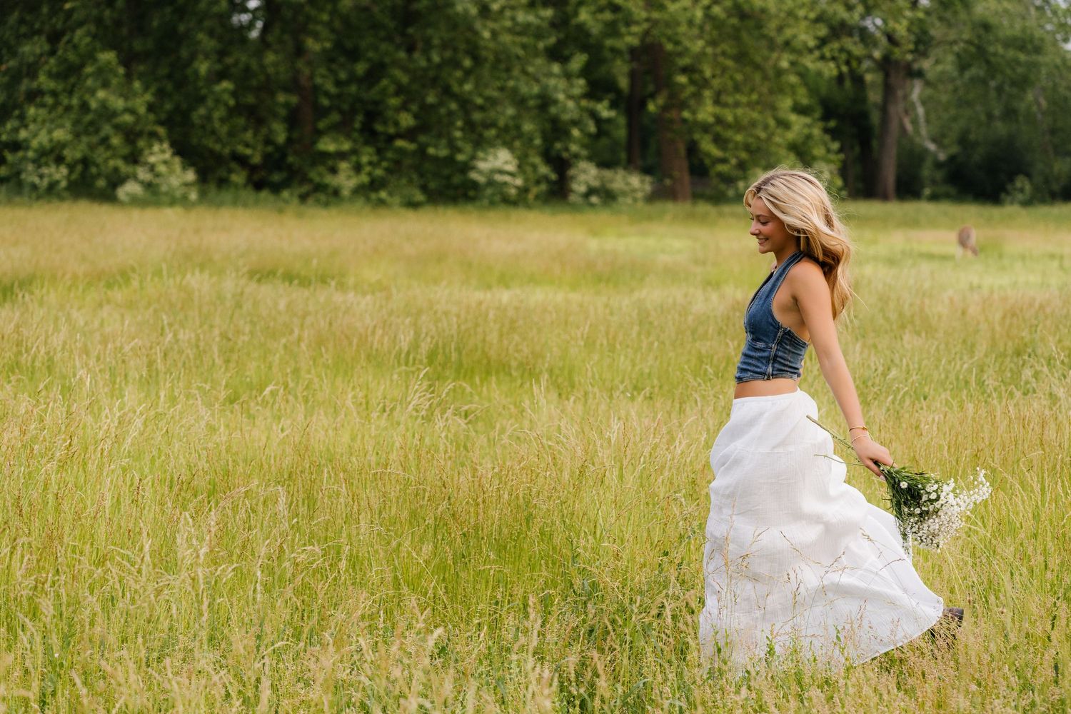 A bride in a flowing white dress walks through a tall grass meadow holding a wildflower bouquet on a sunny summer day.
